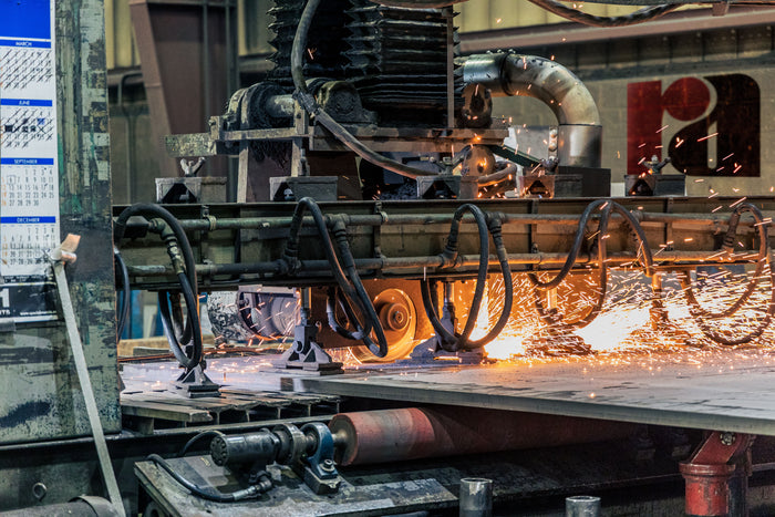 Industrial metal cutting machinery in a factory setting, with bright orange sparks flying as a large steel plate is being processed.